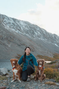 asian girl crouching down with her two sitting dogs. there is a mountain with snow in the background