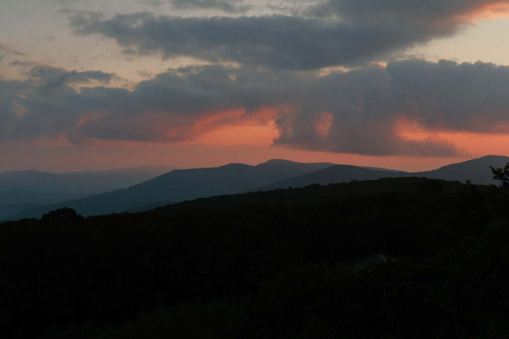 A landscape photo of the blue ridge mountains at sunrise. The sky has dark blue clouds and pink skies. The mountains are shades of gray, black, and blue.